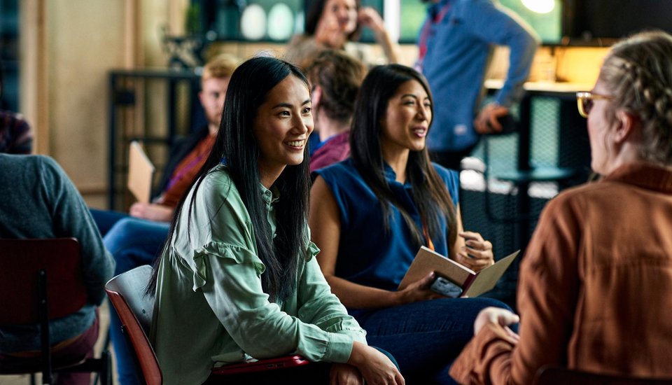 A group of people sit in a circle indoors, smiling and talking. Two women in the foreground are engaged in conversation, one holding a notebook. The setting appears informal and friendly.