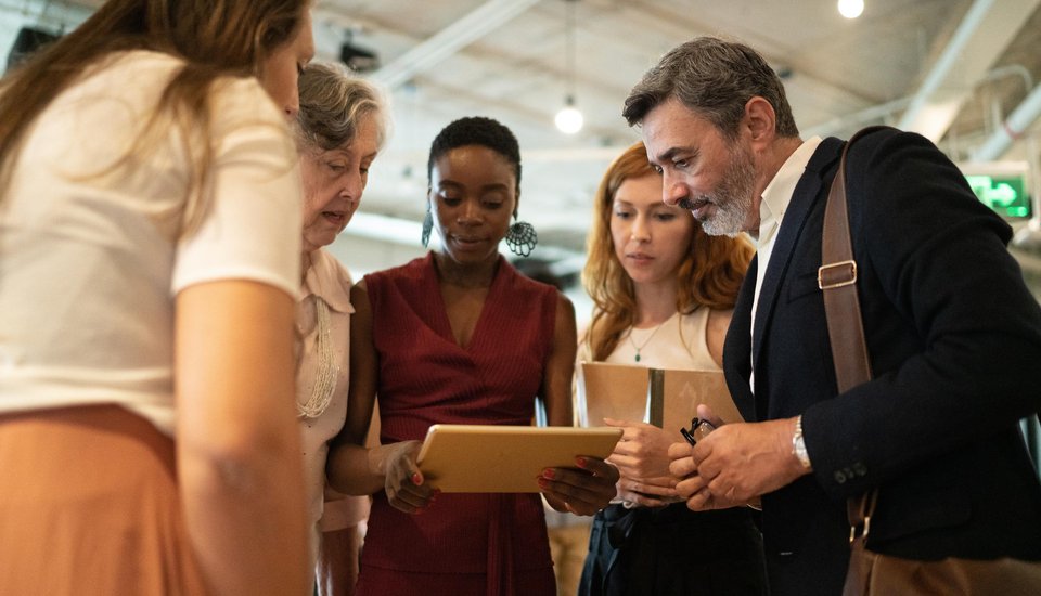 Five people gather closely around a tablet in a modern office, engaged and focused as they collaborate during a creative workshop, exchanging ideas and inspiration as a team.