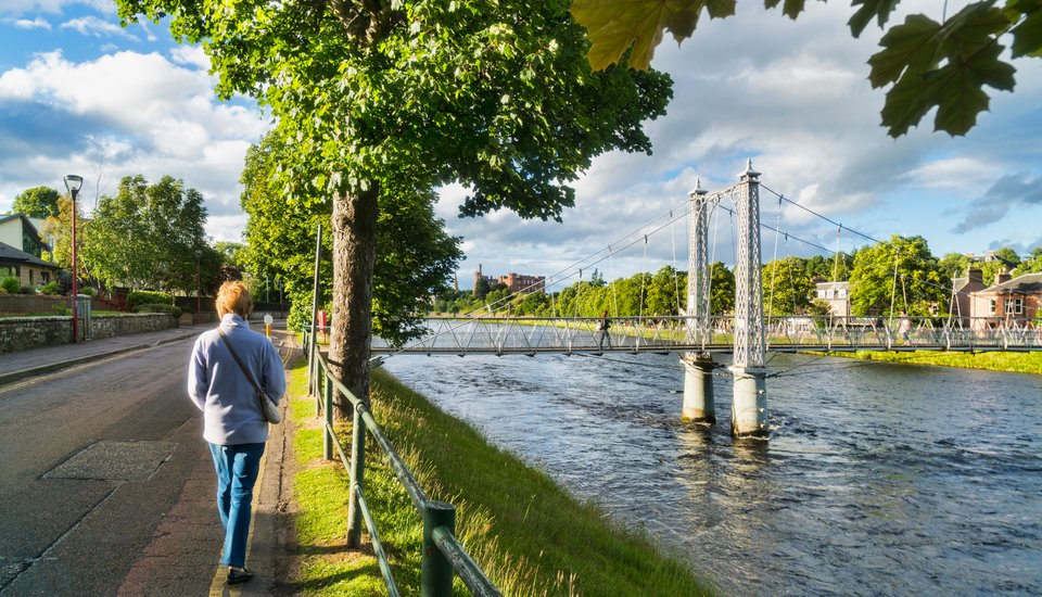 A person walks along a riverside path lined with trees, beside a metal suspension footbridge crossing the river under a partly cloudy sky. Buildings and greenery are visible on both sides of the river.