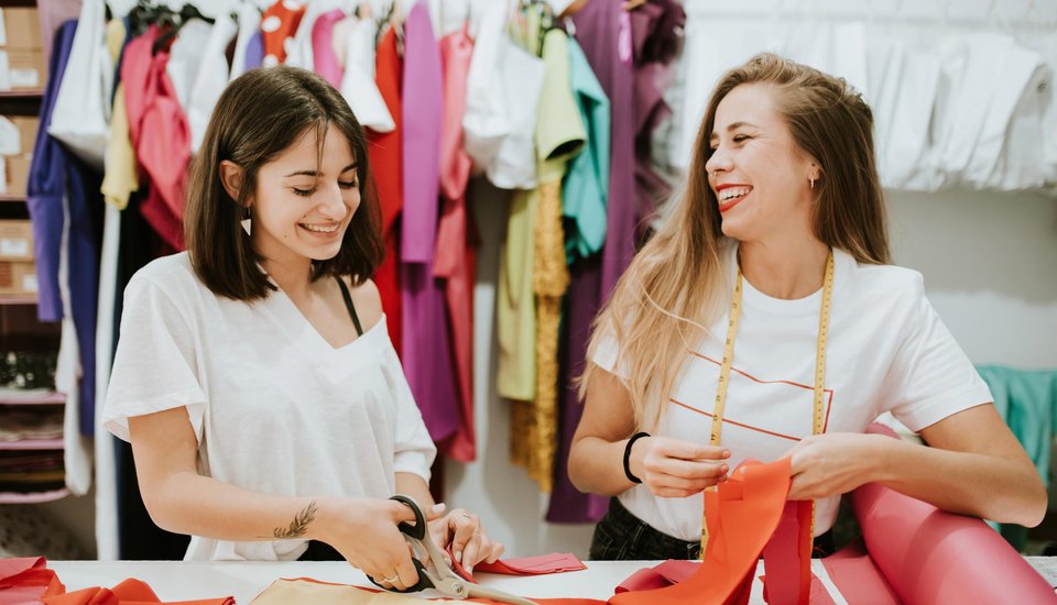 Two women smile while working with fabrics in a colorful sewing studio. One cuts fabric, scissors in hand, while the other holds a measuring tape. The lively space evokes the spirit of a creative workshop, with textiles and clothing hanging in the background.