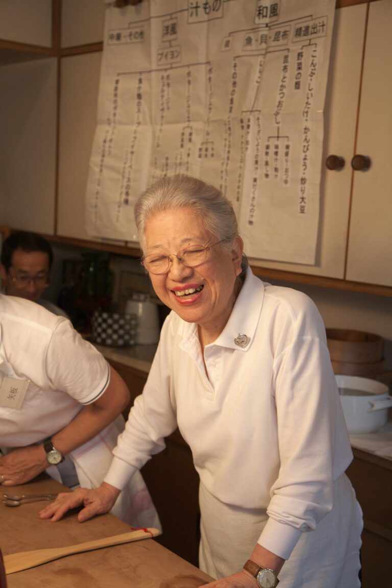 An elderly woman with gray hair and glasses, wearing a white shirt, smiles warmly while leaning on a table in a kitchen. A large handwritten chart is hanging on the wall behind her.