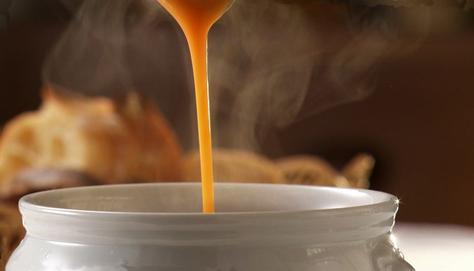A thick, steaming orange soup is being poured into a white bowl, with a blurred background featuring bread.