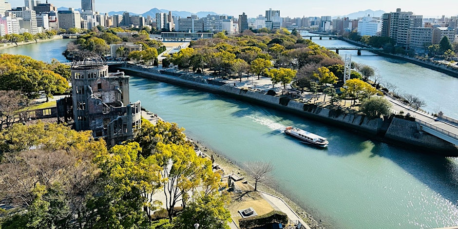 Aerial view of Hiroshima Peace Memorial Park with the Atomic Bomb Dome, river, trees, a passing boat, and surrounding city buildings under a clear sky.