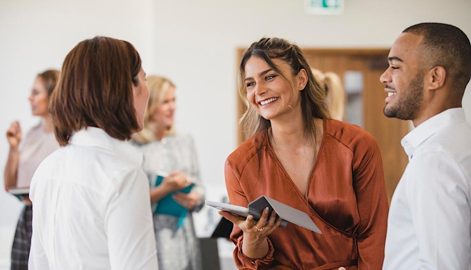 A group of professionals are talking and smiling at a networking event; one woman holds a tablet while engaging with two colleagues, with others chatting in the background.