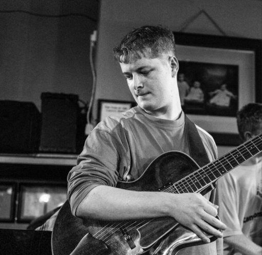 A young man in a light-colored shirt holds an acoustic guitar, looking down thoughtfully. He is indoors, and black-and-white framed photos hang on the wall behind him. The image is in black and white.
