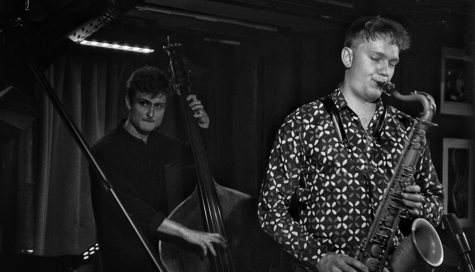 Black and white photo of two musicians performing; one plays an upright bass while the other, in a patterned shirt, plays a saxophone. The background shows dark curtains and dim lighting, suggesting a jazz club setting.