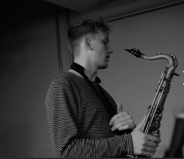 A young man wearing a striped shirt stands in profile, holding a saxophone, with a saxophone strap around his neck. The photo is black and white and appears to be taken indoors.
