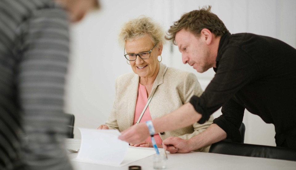An older woman with glasses smiles while looking at papers on a table, as a man beside her points at the documents. A blurred person is in the foreground. They appear to be in a meeting or discussion.