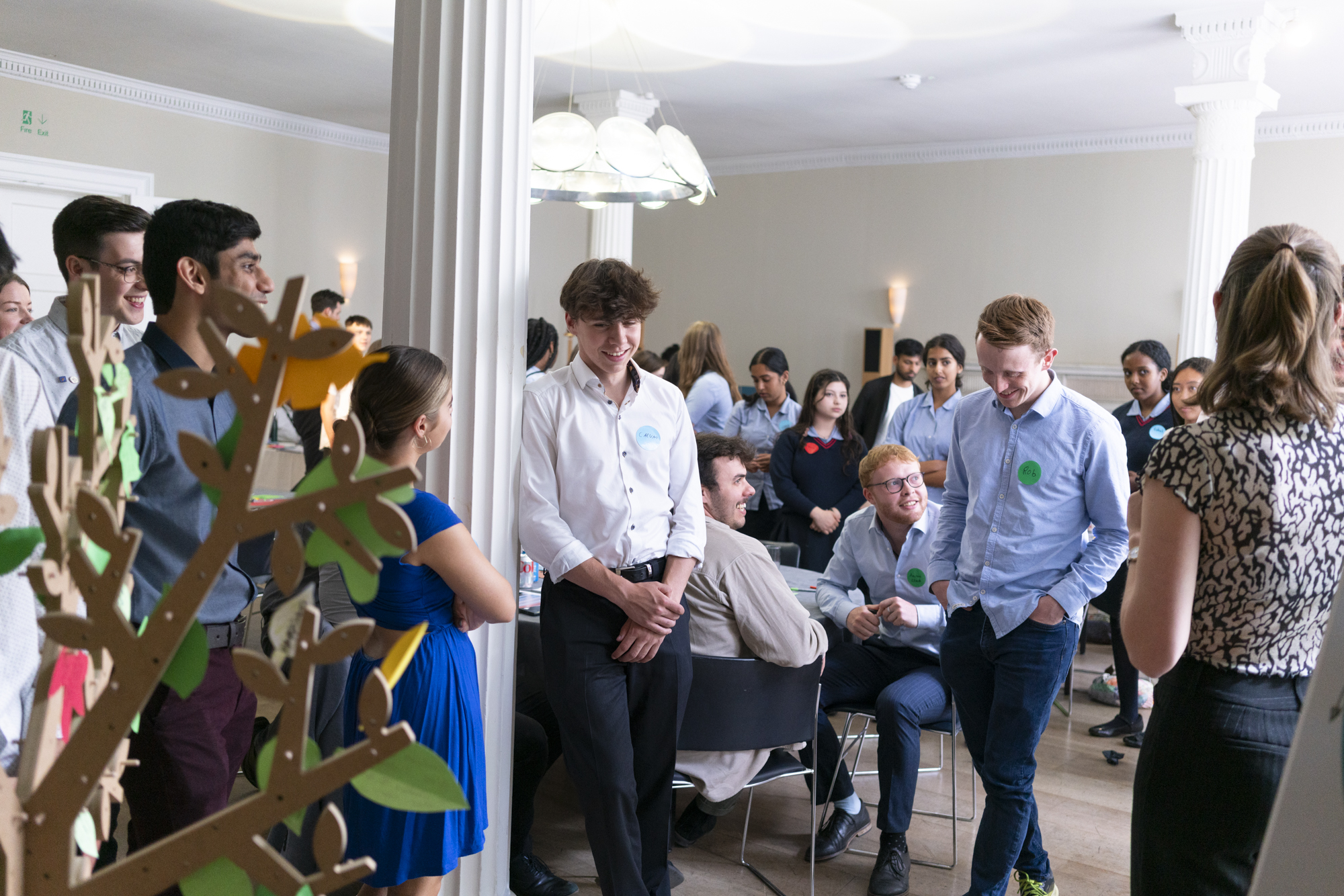 A group of people, mostly young adults, are socializing and conversing in a brightly lit room with white columns, possibly celebrating student design awards. Some stand and smile, while others sit at tables near a decorative tree in the foreground.