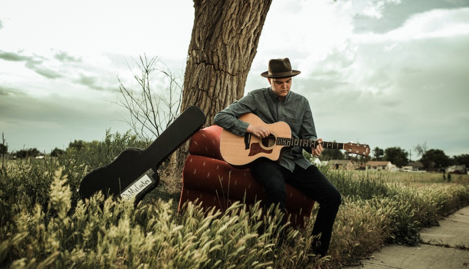 A man in a hat sits on a red couch outdoors, playing an acoustic guitar beside a tree. A guitar case rests in the grass nearby, with a cloudy sky and field in the background.
