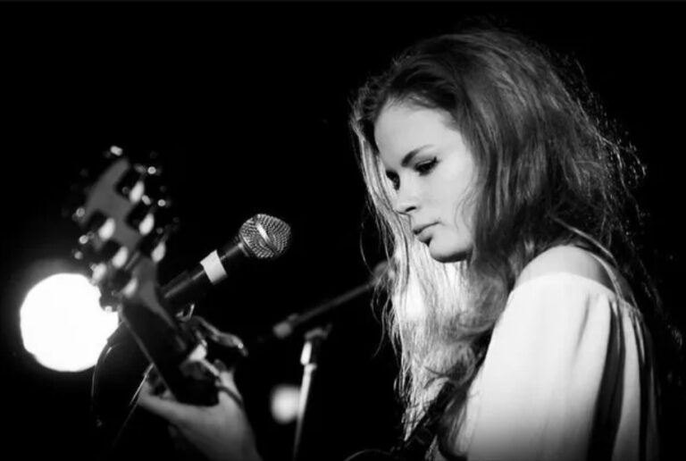 A woman with long hair plays guitar and sings into a microphone on stage. The image is in black and white, with a bright light in the background, creating a moody, focused atmosphere.