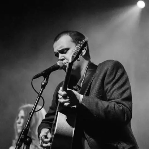 A man in a suit jacket plays an acoustic guitar and sings into a microphone on stage, with a woman visible in the background and dramatic stage lighting.