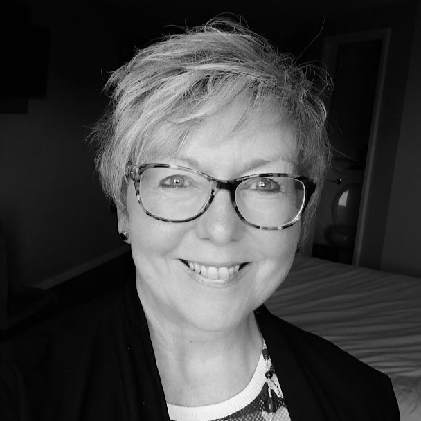 A smiling woman with short, light hair and glasses takes a selfie in a room, possibly before the Edward Boyle Lecture. She is wearing a dark top and patterned shirt underneath. The photo is black and white.