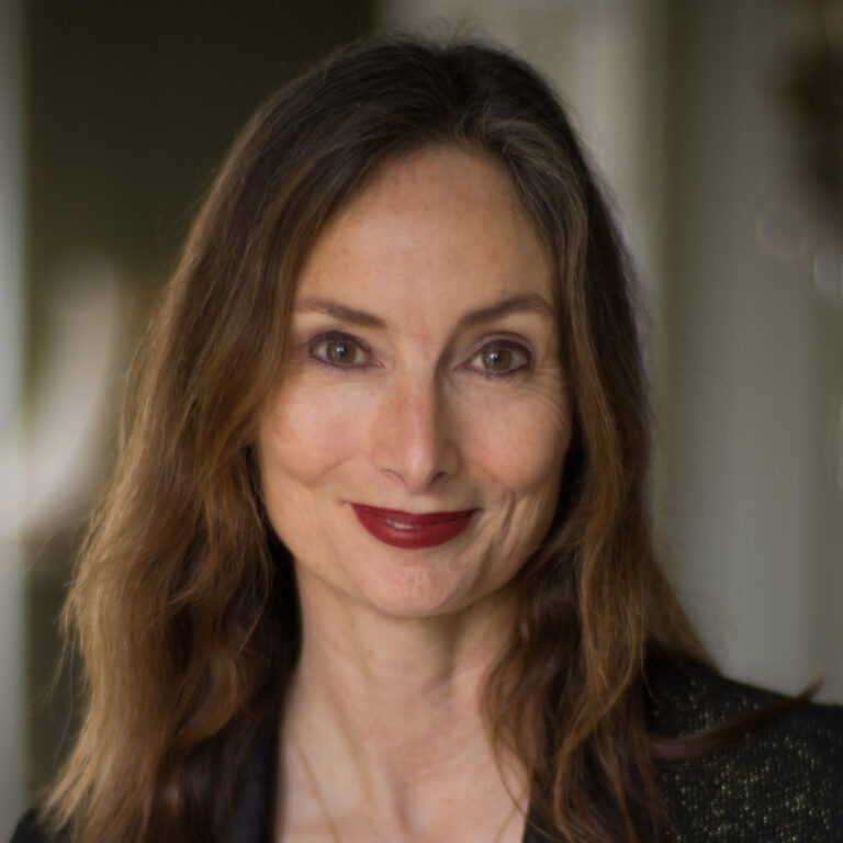 A woman with long brown hair and red lipstick smiles softly at the camera. She is wearing a dark top and the background is softly blurred.