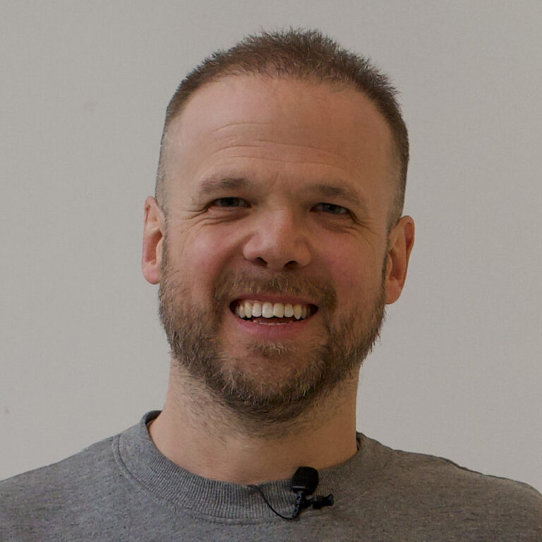 A smiling man with short hair and a beard, wearing a grey shirt and a small microphone clipped to his collar, stands against a plain light-colored background.
