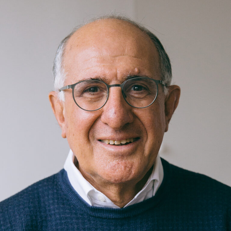 An older man with glasses and short gray hair smiles at the camera. He is wearing a dark blue sweater over a white collared shirt, standing in front of a plain light-colored background.