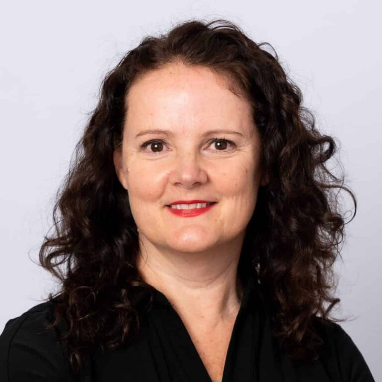 A woman with fair skin and curly dark brown hair is smiling slightly. She is wearing a black top and red lipstick, posing in front of a plain light gray background.