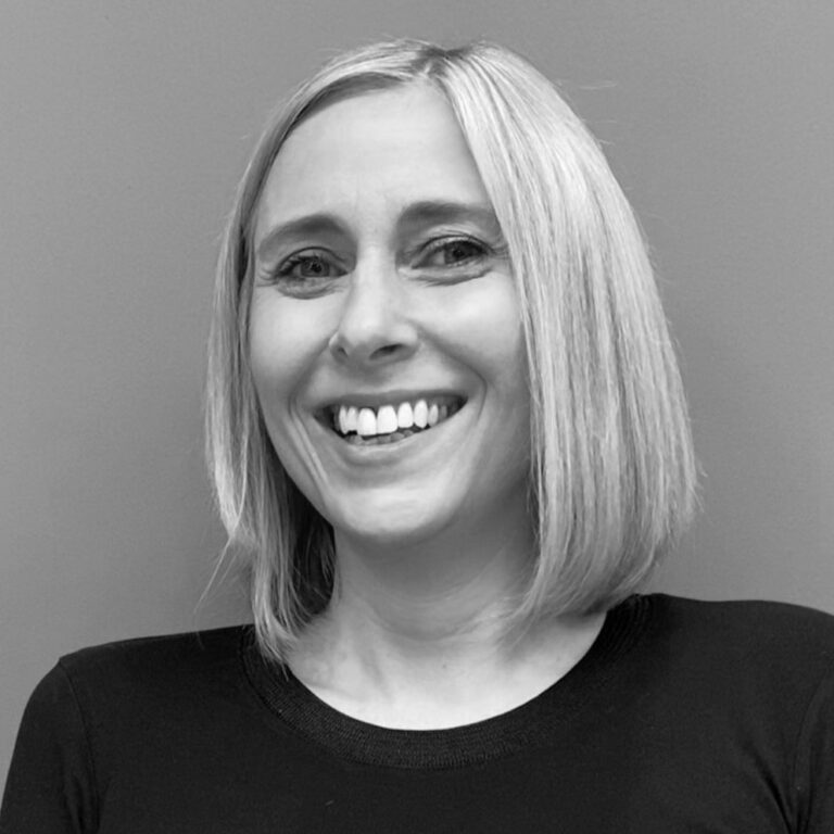 A woman with straight, shoulder-length blonde hair is smiling at the camera. Wearing a dark long-sleeved top, she stands before a plain background in this black and white image highlighting professionals in digital badging.
