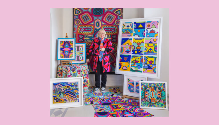 Lisa Todd in a colorful coat stands in a bright room surrounded by her artwork: vibrant, abstract paintings and framed art pieces with bold patterns and colors. The background and floor display more vivid artworks.