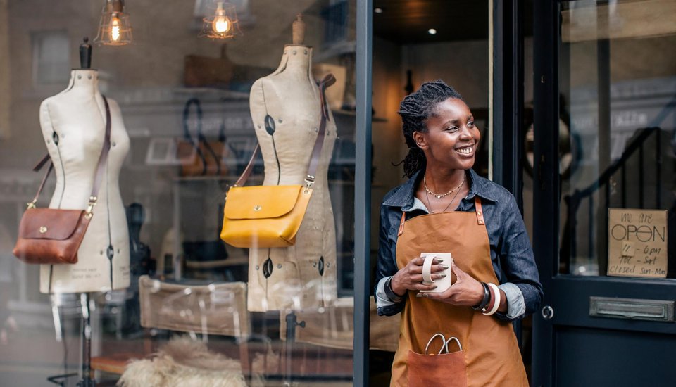 A smiling shop owner in an apron stands at her store entrance holding a coffee mug. Behind her, mannequins display handbags in the window and a handwritten "OPEN" sign hangs on the door, ready to welcome customers attending Newcastle events nearby.