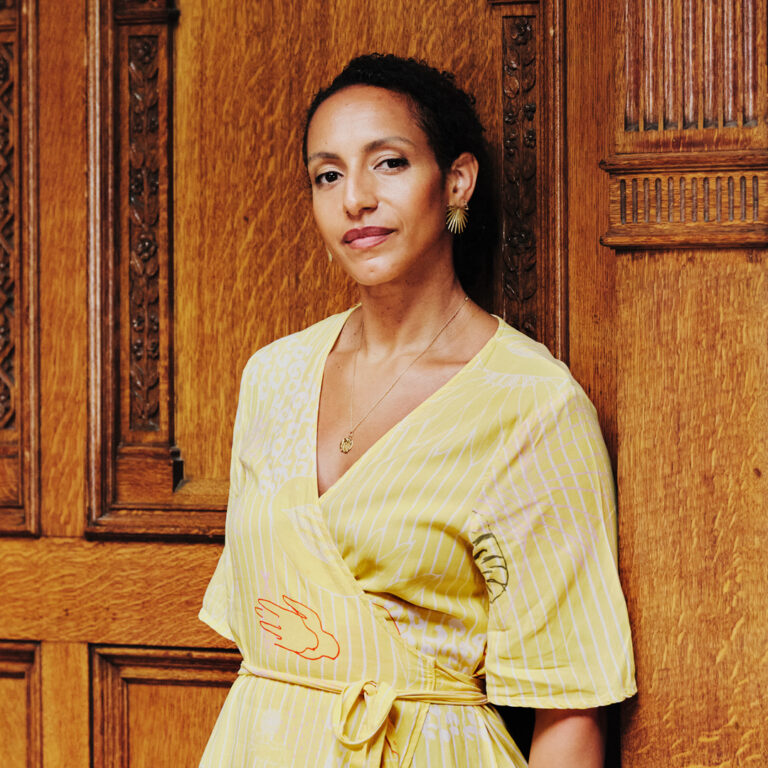 Afua Hirsch with curly hair pulled back, wearing a yellow wrap dress with a hand pattern, stands in front of an ornately carved wooden wall, looking calmly at the camera.