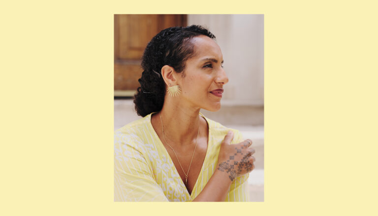 A woman with curly dark hair wearing a yellow patterned top, gold sunburst earrings, and a necklace looks to the side. She has a henna design on her hand and stands in front of a blurred wooden door and light background.