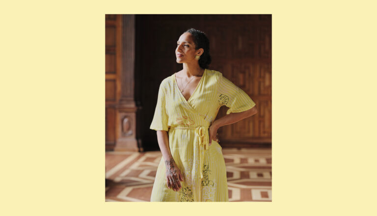 A woman in a yellow wrap dress stands indoors on a patterned wooden floor, gazing to the side with a slight smile. She has dark hair tied back and is surrounded by wood-paneled walls.