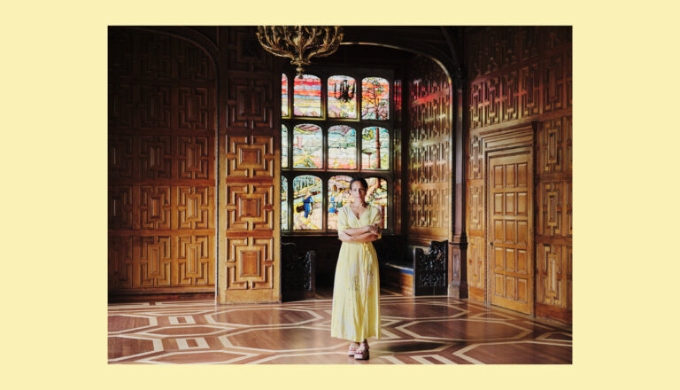 A woman in a yellow dress stands in the center of an ornate wooden room with geometric parquet floors and colorful stained glass windows, illuminated by natural light.
