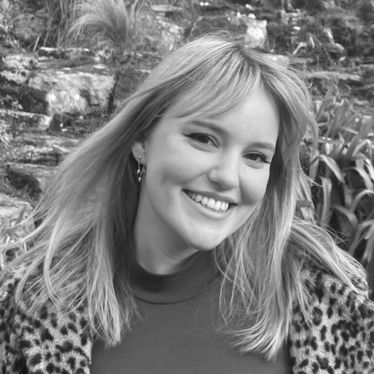 A young woman with straight, light hair smiles at the camera. She is wearing a patterned jacket and standing outdoors with plants and rocks in the background. The photo is in black and white, capturing the spirit of student design awards creativity.