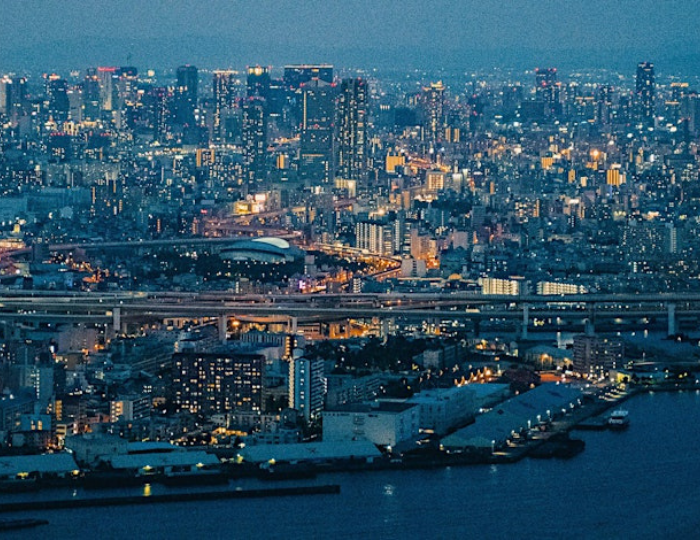 Aerial view of a cityscape at dusk with buildings illuminated, roads, and a body of water in the foreground, capturing a vibrant urban night scene reminiscent of the energy surrounding the Edward Boyle Lecture.