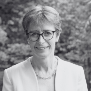 A woman with short hair and glasses, wearing a light-colored blazer and necklace, smiles at the camera. The background is blurred greenery, reflecting the atmosphere of a sustainability event. The image is in black and white.