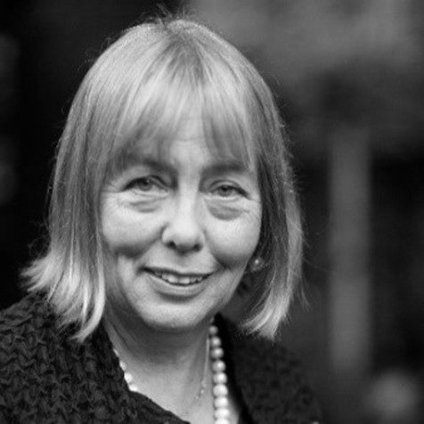 A black-and-white portrait of an older woman with light-colored hair, wearing a textured dark top and a pearl necklace, smiling softly at the camera. The blurred background hints at her East Midlands roots and commitment to inclusive growth.