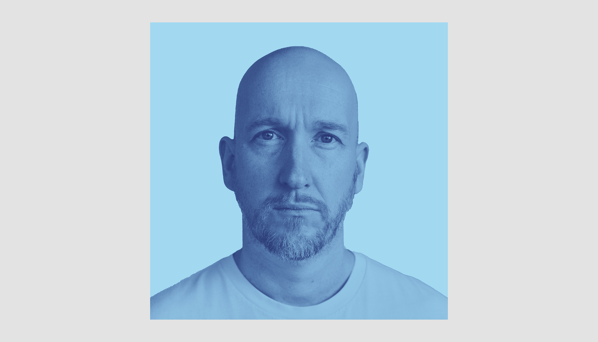 A bald man with a trimmed beard and mustache looks directly at the camera with a neutral expression. He is wearing a plain white t-shirt, and the background is a solid light blue color.