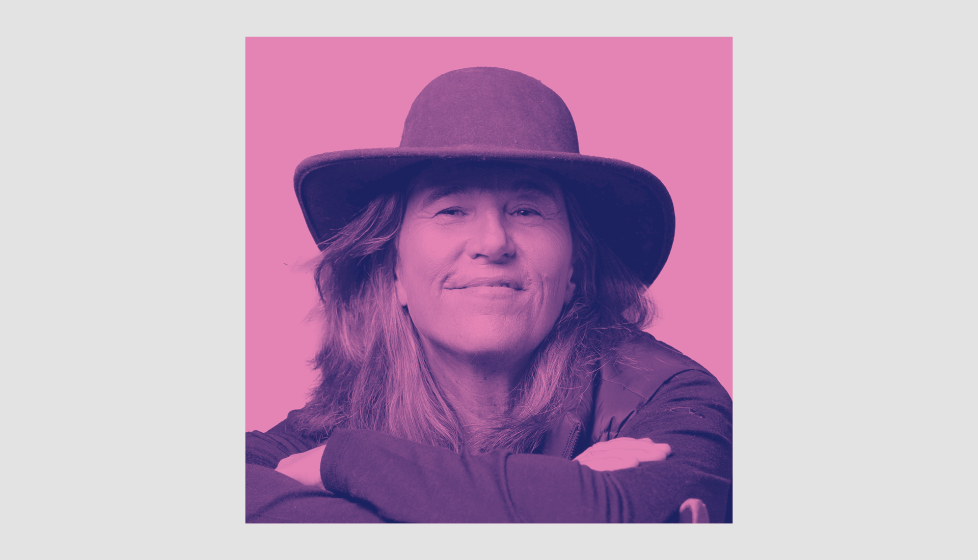 A person with long hair and a wide-brimmed hat smiles gently, resting their arms on a surface. The image has a pink and purple color filter.