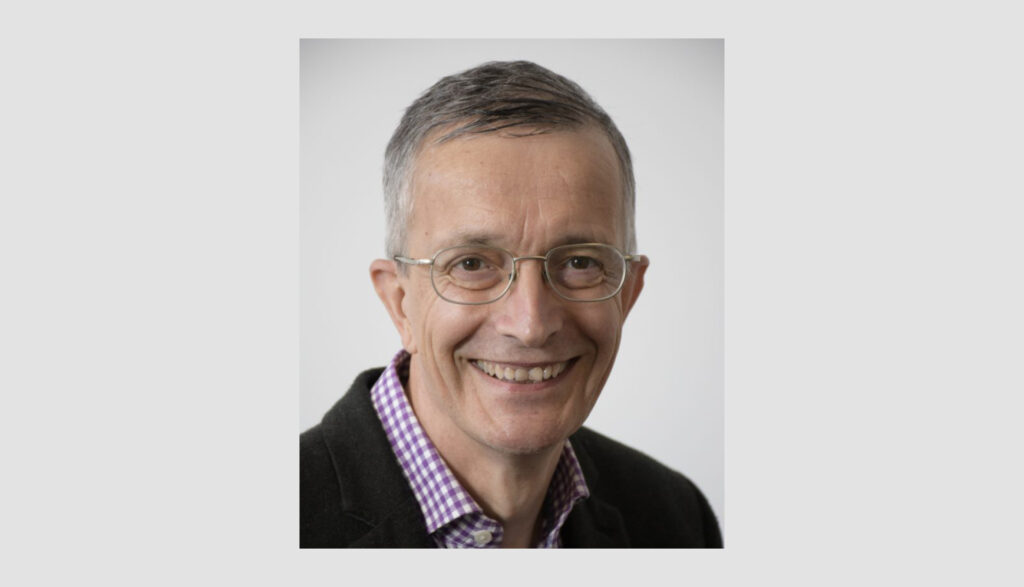 A smiling older man with short gray hair, wearing glasses, a dark blazer, and a purple checkered shirt, in front of a plain light gray background.