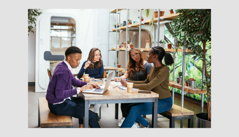 Four people sit around a table in a modern office, talking and working on laptops with coffee cups nearby. The setting features large windows, plants, and a bright, collaborative atmosphere.