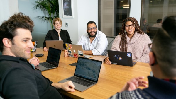 Five people sit around a conference table with laptops, smiling and engaged in discussion as they connect and build community in a modern office setting. A plant and large windows are visible in the background.