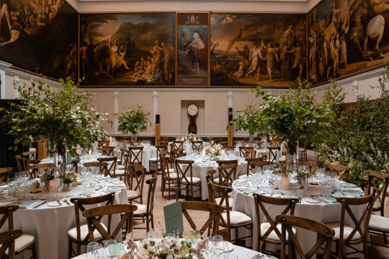 Elegant banquet hall with large, classical wall paintings, round tables set with white tablecloths, glassware, and floral centerpieces, surrounded by wooden chairs and decorated with lush greenery.
