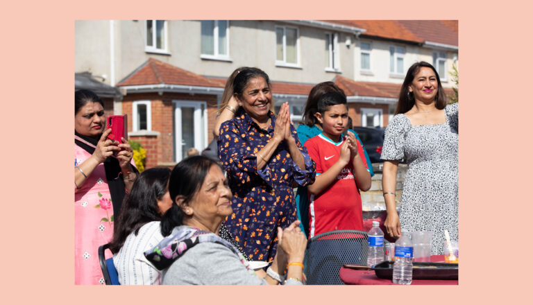 A group of people, including women and a boy, stand and sit outdoors in sunlight, some clapping and one holding a phone to take a photo. Houses and a table with water bottles are visible in the background.