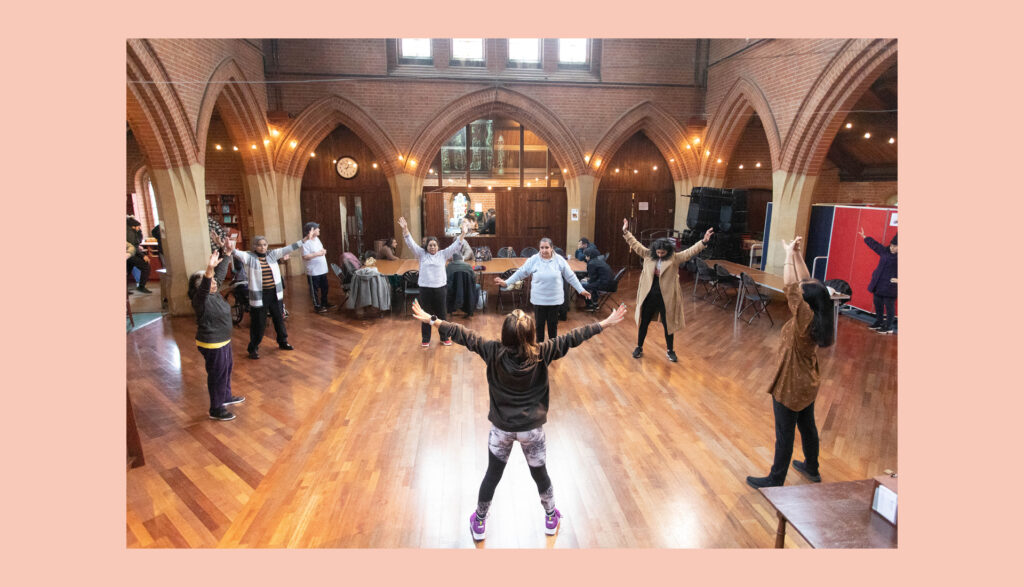 A group of people stand in a circle with arms outstretched, participating in an activity inside a spacious hall with arched windows, wooden floors, and warm lighting.