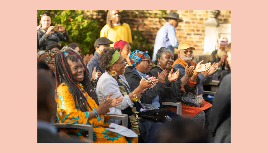 A group of people sitting outdoors, smiling and clapping, enjoying an event. They are dressed in colorful clothing and head wraps, with trees and a brick wall in the background.