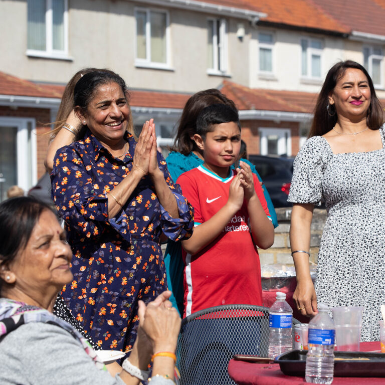 A group of people, including women and a boy in a red shirt, stand and sit outdoors, smiling and clapping, with houses in the background and a table with drinks in the foreground.