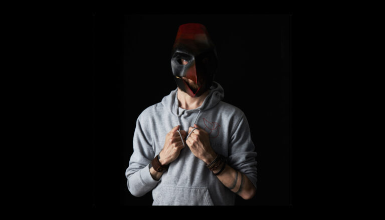 A person wearing a gray hoodie and leather bracelets stands against a black background, holding the hoodie strings, and wearing a dark, bird-like mask with red and black colors.