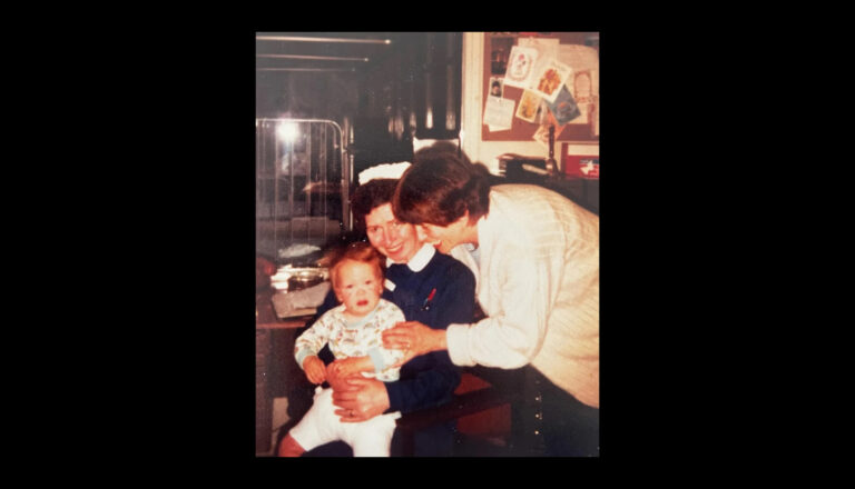 A nurse in uniform holds a crying baby on her lap while another woman in a sweater leans in, smiling, in a warmly lit room decorated with cards and photos.