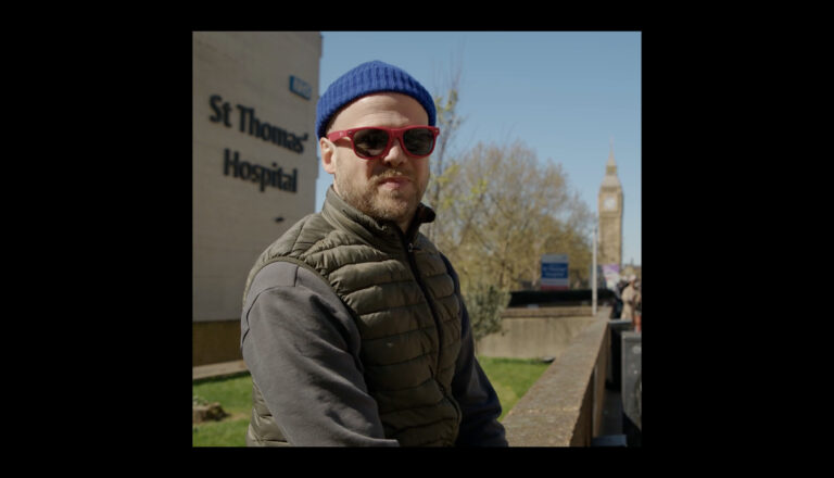 A man in a blue beanie, red sunglasses, and a padded vest sits outside St Thomas' Hospital with the Big Ben clock tower visible in the background under a clear blue sky.
