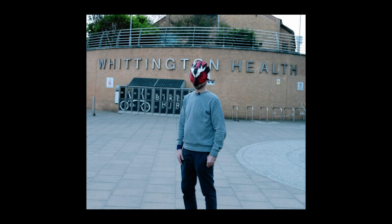 A person wearing a red and white mask stands outside Whittington Health, with bike racks and a brick building visible in the background.