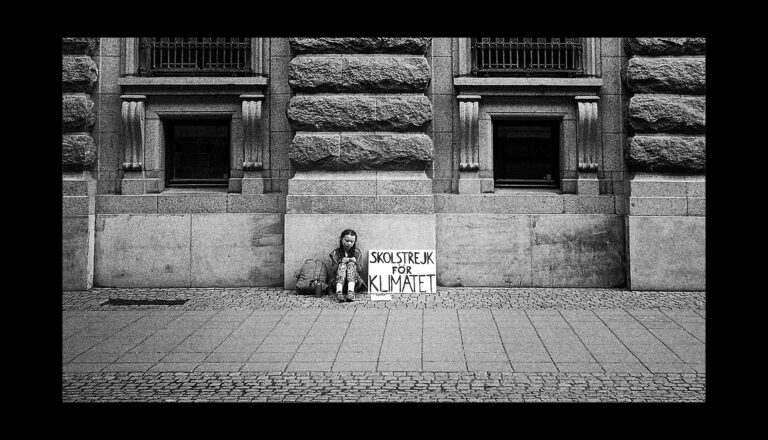 A young person sits alone on a sidewalk against a stone building, holding a sign that reads "Skolstrejk för klimatet" ("School strike for climate") in Swedish. Two bags are placed on either side of them.