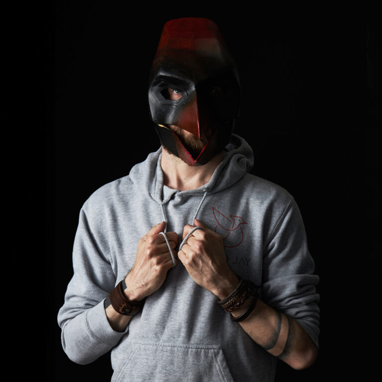 A person wearing a dark, bird-shaped mask and a gray hoodie stands against a black background, holding the hoodie’s collar with both hands. The person has bracelets on each wrist and a bird emblem on the hoodie.