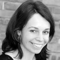 A woman with shoulder-length dark hair smiles at the camera during the Edward Boyle Lecture. She is wearing a light-colored top and small hoop earrings. The background is blurred, showing a brick wall. The image is in black and white.