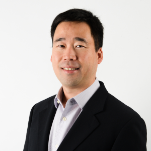 A man with short black hair wearing a black blazer and light-colored shirt, smiling slightly, stands against a plain white background, ready to speak at a sustainability event focused on the UN Sustainable Development Goals.